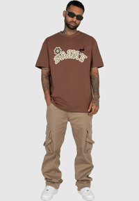 Brun bomulds T-shirt med fed cremefarvet tekst og grafiske designs; parret med lysebrune cargo bukser og hvide sneakers.
