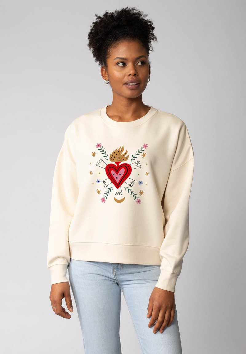 Vrouw met krullend haar draagt een crèmekleurige sweatshirt met een rode hart, vlammen, bloemen, sterren en vleugelillustraties, in combinatie met lichtblauwe jeans.