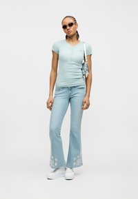 Γυναίκα με ανοιχτό γαλάζιο εφαρμοστό πουκάμισο με κουμπιά, καμπάνα τζιν με λευκή λουλουδάτη κεντητική, λευκά αθλητικά παπούτσια, γυαλιά ηλίου και ασημένια τσάντα ώμου.