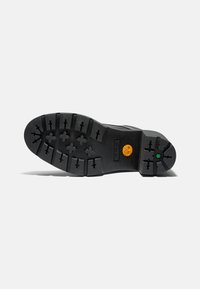 Suela de goma negra con surcos profundos, marca visible y acentos de color. Presenta un tacón ancho y plano y una superficie texturizada para mejorar el agarre.