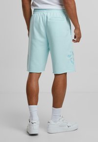 Lyseblå bomullsblandingsshorts med elastisk midje, sidelomme og dekorativt broderimønster nederst til venstre.