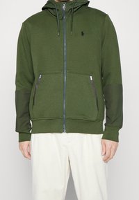 Grön zip-up hoodie med en dragsko på huva, med två fickor framtill med dragkedja och svarta detaljer på de nedre ärmarna.