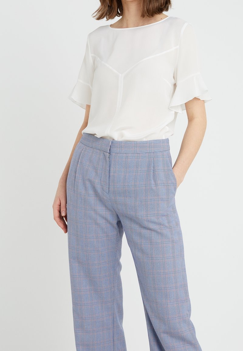 Weiße Chiffonbluse mit Rüschenärmeln, kombiniert mit hellblauen karierten Hosen mit einem dezenten Karomuster und seitlichen Taschen.