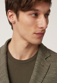 Olijfgroene blazer van textuurstof, vergezeld van een strakke olijfgroene trui. De blazer heeft subtiele gouden reversspelddetails.
