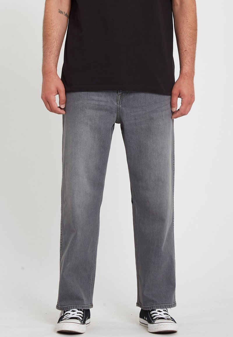 Vaqueros grises de denim con un ajuste relajado, cinco bolsillos y un desgaste sutil. Combinados con zapatillas negras y una camiseta negra.