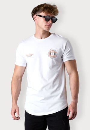 T-shirt blanc à manches courtes avec un graphique de soleil en orange et noir accompagné de texte, assorti de lunettes de soleil noires et d'un jean noir déchiré.