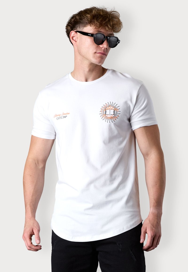 T-shirt blanc à manches courtes avec un graphique de soleil en orange et noir accompagné de texte, assorti de lunettes de soleil noires et d'un jean noir déchiré.