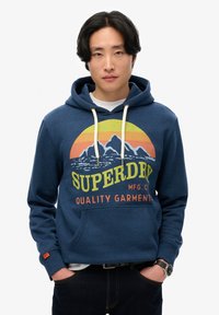 Sudadera azul marino con un gráfico circular que presenta montañas y colores de atardecer, logo de 'SUPERDRY', cordones y un bolsillo frontal.