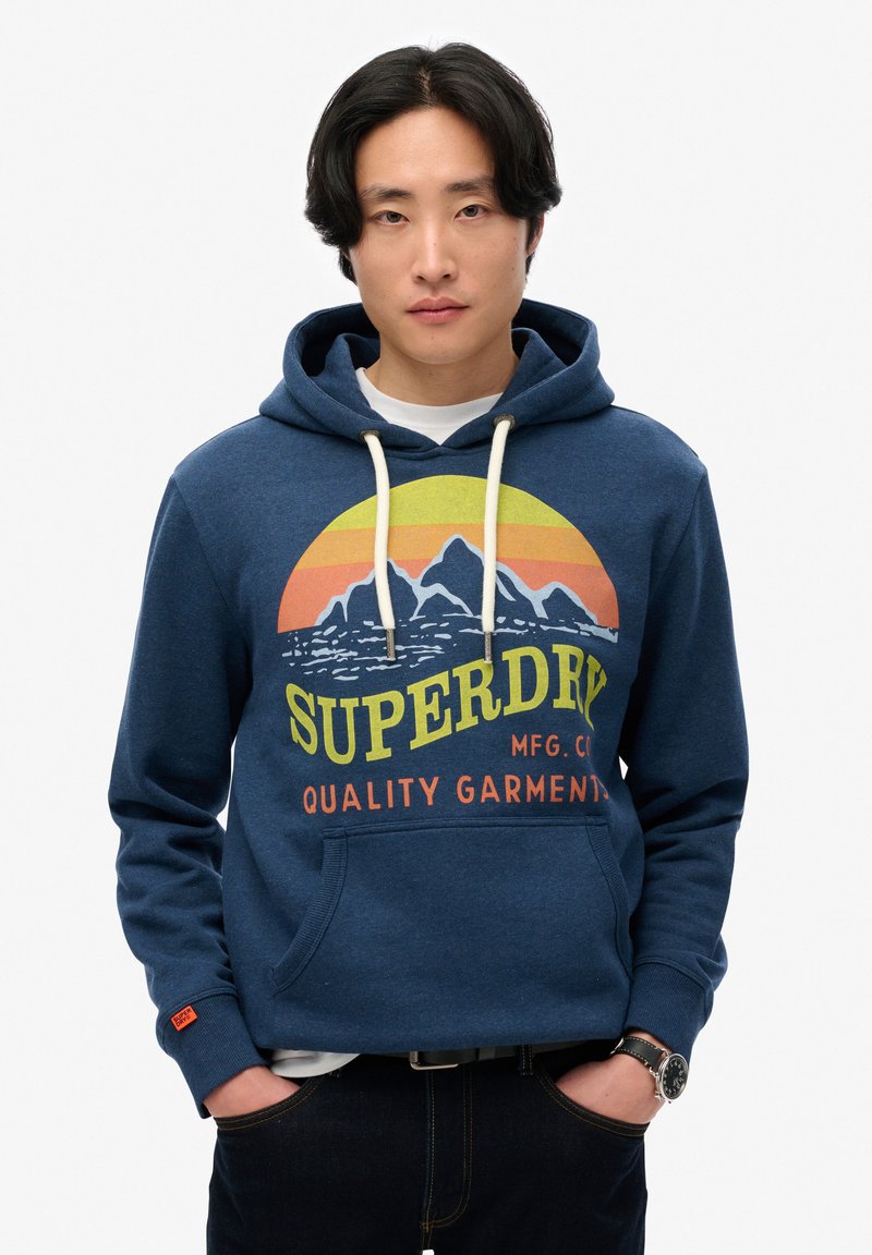 Sudadera azul marino con un gráfico circular que presenta montañas y colores de atardecer, logo de 'SUPERDRY', cordones y un bolsillo frontal.