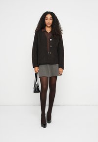 Zwarte wolmix jas met een boxy snit, voorzien van drie knopen, samen met een bruine top, grijze rok en zwarte accessoires.