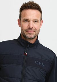Navy Zip-Jacke mit durchgehendem Reißverschluss, bestehend aus einem gesteppten Vorderteil, glatten Ärmeln und einem Logo "FQ1924". Das Material scheint leicht und strukturiert zu sein.