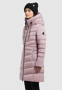 Kvinde iført en lyserød quilteret pufferjakke med hætte og sort beanie, der står i profil mod en ensfarvet baggrund.