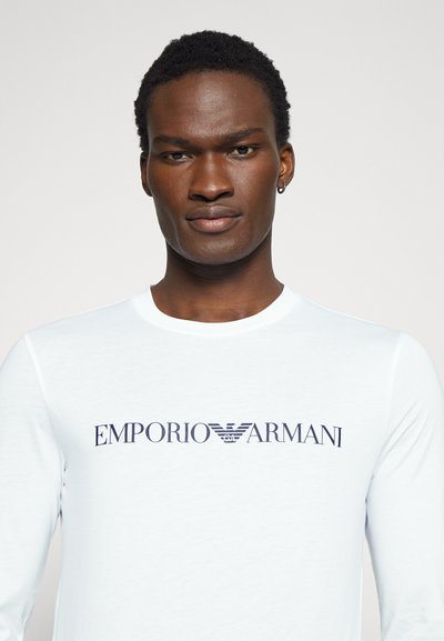 Hvit langermet skjorte laget av myk stoff, med en mørkeblå "EMPORIO ARMANI" logo over brystet i en fet, moderne skrifttype.