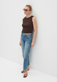 Bruine mouwloze top gecombineerd met lichtblauwe straight-leg jeans en zwarte slippers. De persoon leunt tegen een witte muur.
