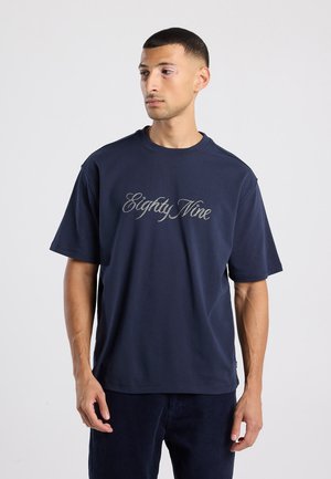 Marineblauwe katoenen T-shirt met korte mouwen en een ronde hals. Bevat de tekst "Eighty Nine" in een zilveren, cursieve lettertype over de voorkant gedrukt.