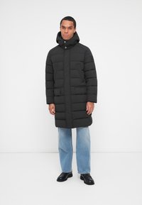 Manteau en duvet noir avec un col haut, fermeture éclair à l'avant et boutons-pression. Doté de deux poches latérales et d'une texture matelassée. Porté par-dessus un jean bleu clair.