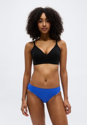 Γυναίκα με σγουρά μαλλιά που φοράει μαύρο σουτιέν και μπλε εσώρουχα, στέκεται μπροστά σε ένα απλό φωτεινό γκρι φόντο.