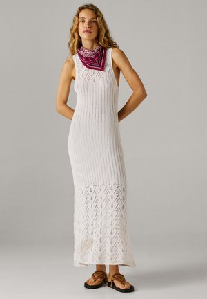 Frau trägt ein ärmelloses, weißes Maxi-Kleid aus Strick mit gemustertem Saum, braune Sandalen und ein lila Bandana um den Hals, steht mit den Händen hinter dem Rücken.