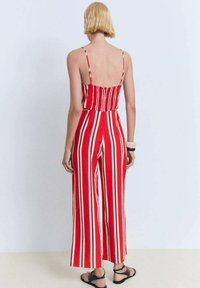 Combinaison rayée rouge et crème avec des fines bretelles, dos smocké, jambes larges et coupe décontractée. Complétée par une simple sandale à bande noire.