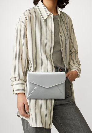 Person trägt ein beige-schwarz gestreiftes Hemd mit Knöpfen, graues Oberteil und graue Jeans mit einer silbernen Umhängetasche im Umschlagstil.