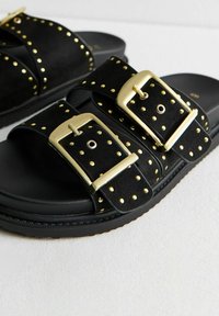 Sandales noires avec deux larges sangles, ornées de clous dorés. Les boucles sont également dorées. Matériau en suede texturé, semelle intérieure rembourrée.
