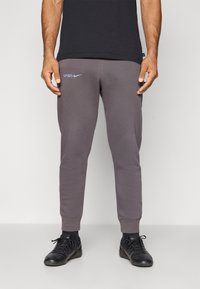Graue Jogginghose mit gerippten Bündchen, versehen mit einem weißen Nike-Logo und dem Text "SPURS" in Hellblau. Hergestellt aus weichem Stoff. Getragen mit dunklen Schuhen.