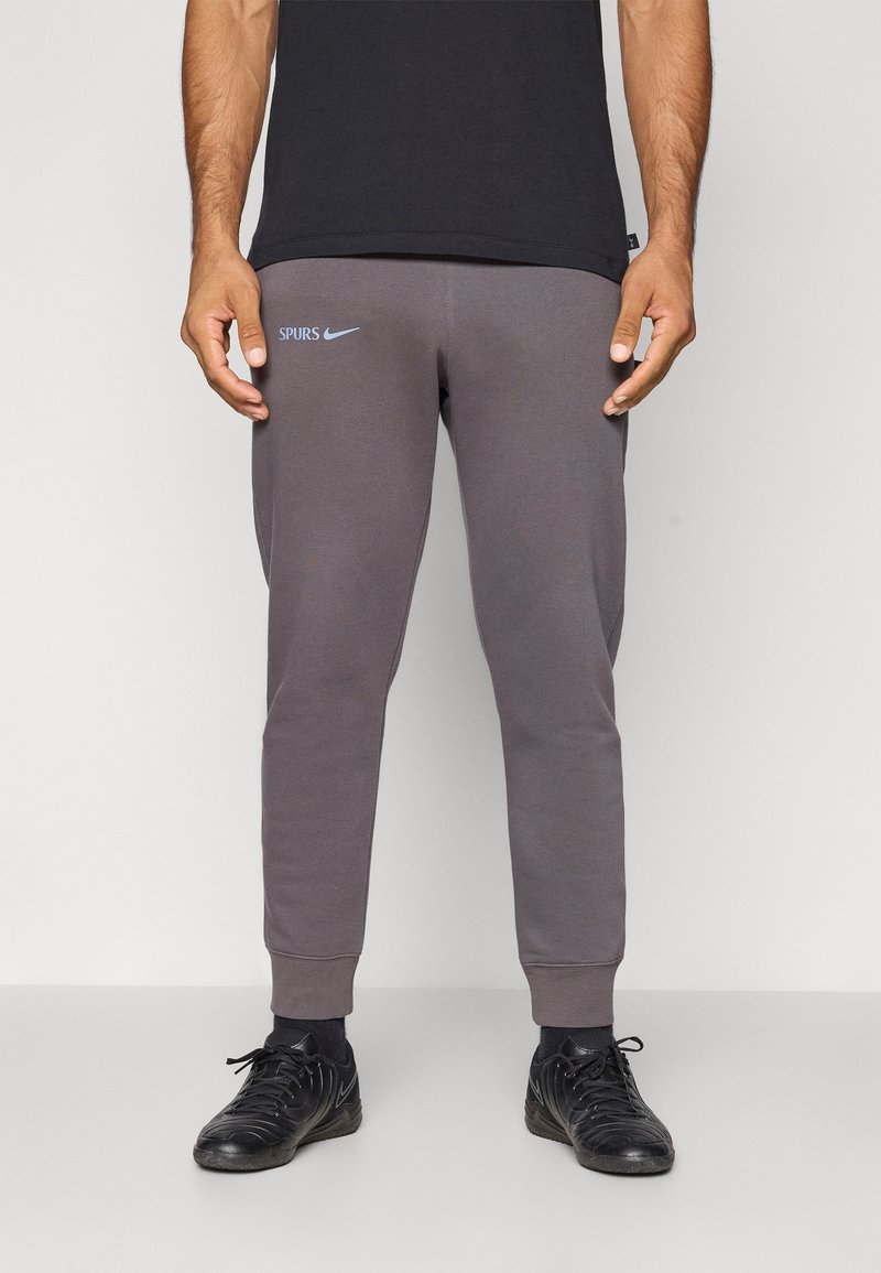 Graue Jogginghose mit gerippten Bündchen, versehen mit einem weißen Nike-Logo und dem Text "SPURS" in Hellblau. Hergestellt aus weichem Stoff. Getragen mit dunklen Schuhen.