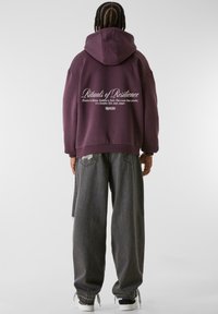 Bordeaux hettegenser med oversized passform, med tekst på ryggen. Kombinert med løse grå jeans og hvite joggesko, som viser en uformell stil.