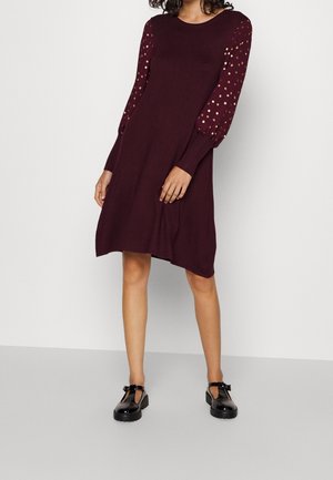 Femme portant une robe bordeaux longueur genoux avec de longues manches parsemées d'or et des chaussures à bride T en cuir verni noir, se tenant sur un sol gris.