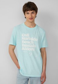 Lichtblauw katoenen T-shirt met grote witte tekst die "Cali Surfside Beach Rentals & More" zegt en schetsen van surfplanken bevat.