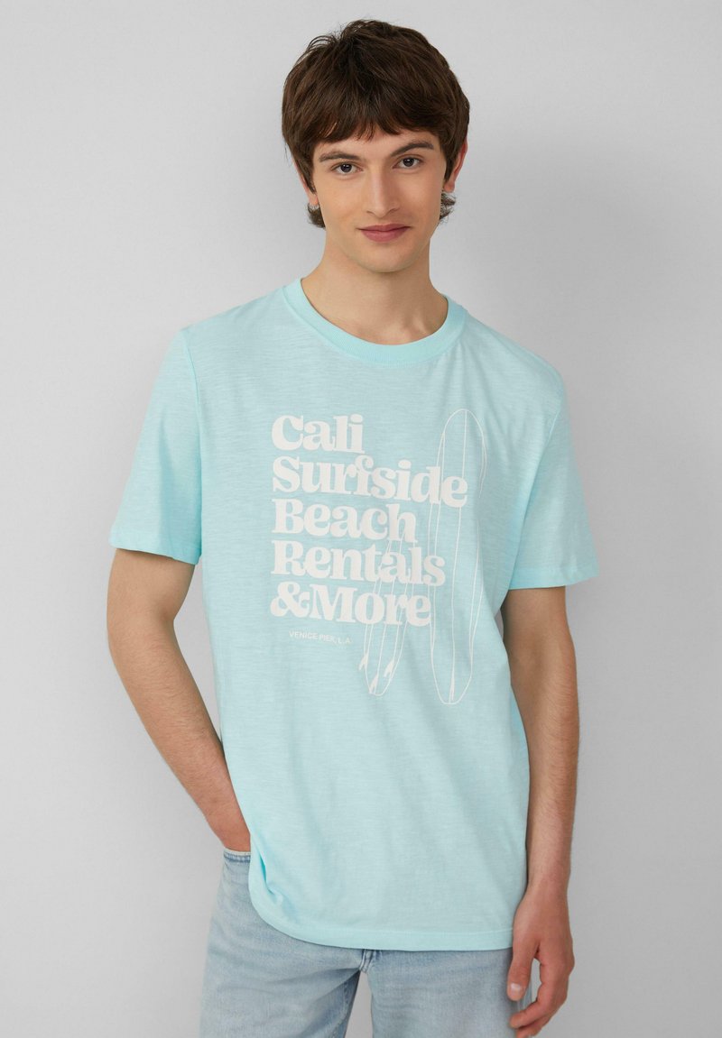 Lichtblauw katoenen T-shirt met grote witte tekst die "Cali Surfside Beach Rentals & More" zegt en schetsen van surfplanken bevat.