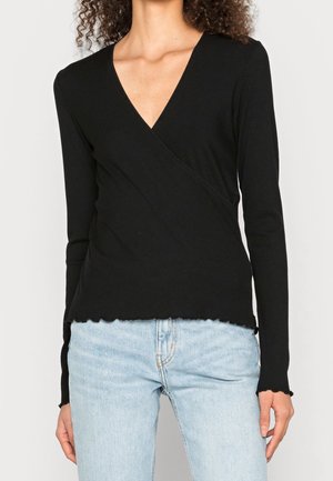 Mujer con una blusa negra de manga larga estilo cruzado con bordes en forma de ondas, combinada con jeans de mezclilla azul claro.