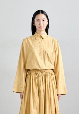 Woman with long black hair wearing gold ball earrings, a loose yellow button-up shirt, and a matching gathered skirt against a light background.