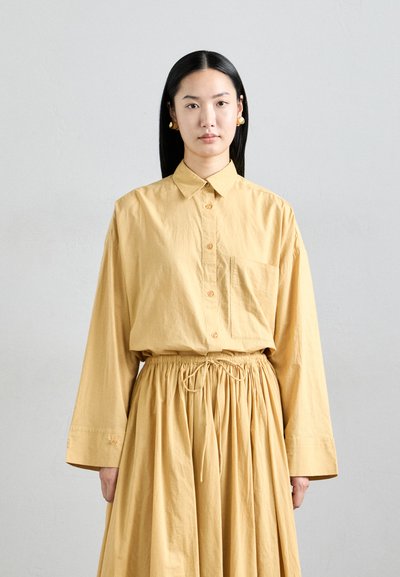 Femme aux longs cheveux noirs portant des boucles d'oreilles en or, une chemise jaune boutonnée ample et une jupe froncée assortie devant un fond clair.