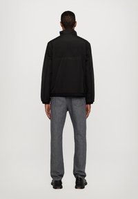 Giacca nera imbottita con collo alto e orlo elasticizzato, con logo tonale sul retro. Indossata con jeans grigi e scarpe nere.