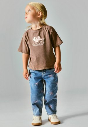 Enfant aux longs cheveux blonds porte un t-shirt marron avec un motif de tasses, un jean bleu avec des nœuds roses et des baskets beiges, regardant en haut à gauche.
