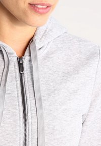 Sweat à capuche gris à fermeture éclair avec un tissu texturé, doté d'une capuche à cordon de serrage et d'une fermeture éclair noire, mettant en avant un design décontracté et une texture douce.