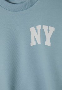 Světle modrá mikina vyrobená z bavlněné tkaniny, která má ribbovaný výstřih a bílý potisk "NY" na přední straně. Hladká textura, ležérní design.