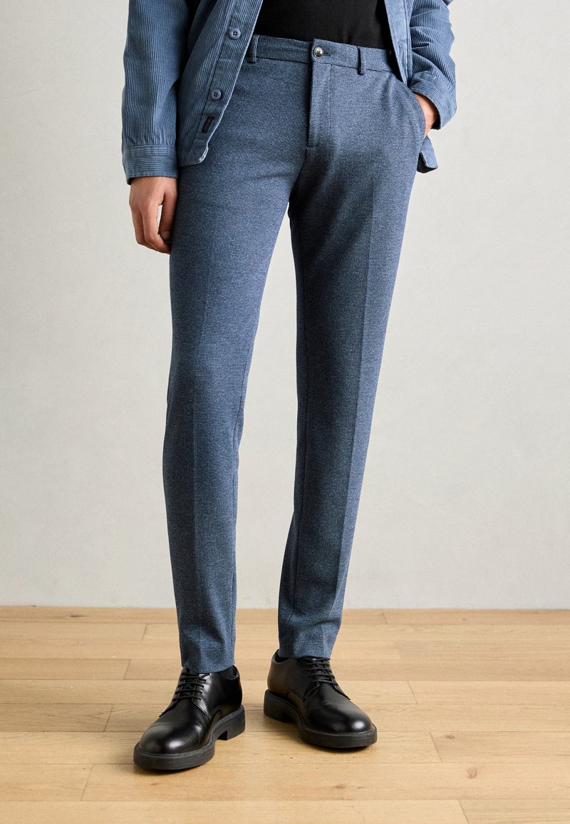 Pantalones azul texturizados con corte ajustado, que presentan un acabado suave, bolsillos frontales y zapatos negros con un diseño de punta redonda.