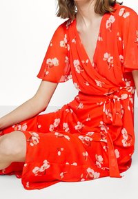 Robe portefeuille rouge à fleurs avec manches courtes, confectionnée en tissu léger. Présente une taille nouée, un ourlet à hauteur du genou et un délicat motif floral.