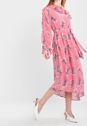 Vrouw in een roze bloemenjurk met één schouder, ruches aan de mouwen en witte slingback hakken, staand tegen een eenvoudige lichte achtergrond.