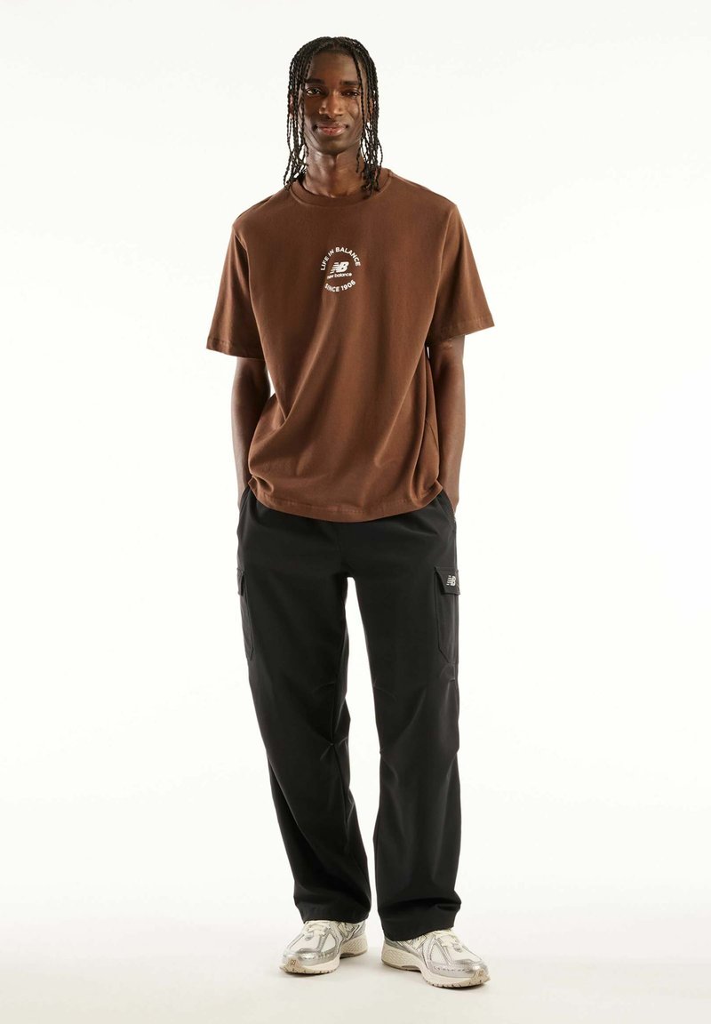 Young man with braided hair stands wearing brown New Balance T-shirt, black cargo pants with logo, and white sneakers against white background.