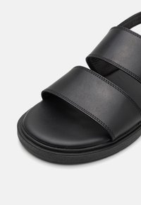 Sorte svart lær sandaler med to brede stropper, teksturert såle og en glatt fotseng, designet for komfort og enkelhet.