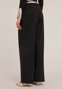Zwarte wijde broek gemaakt van een gladde stof, met een strak ontwerp en subtiele plooien, gecombineerd met lichtgekleurde hakken.