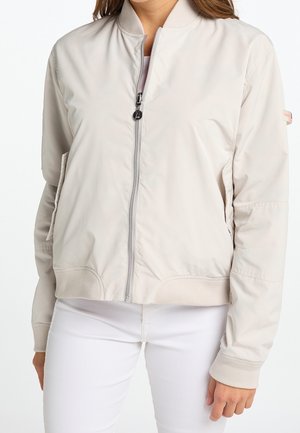 Femme portant une veste bomber beige clair avec fermeture éclair à l'avant, col, poignets et ourlet côtelés, associée à un pantalon blanc.