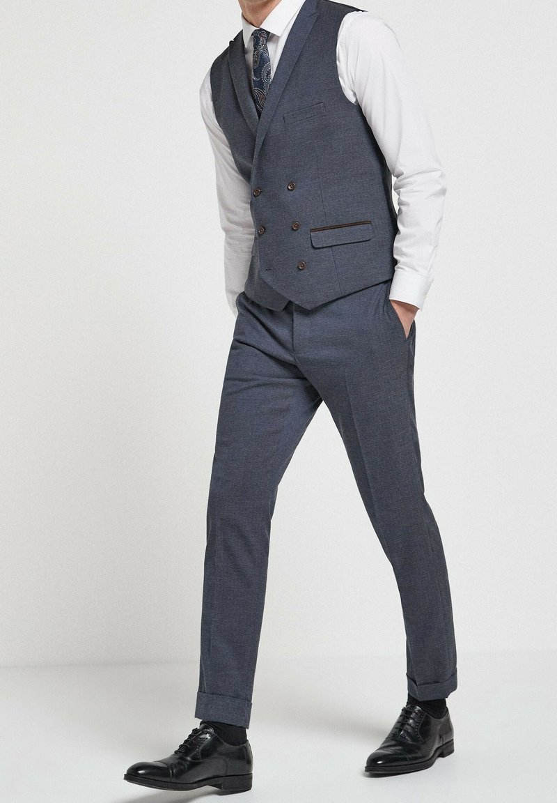 Dopasowany szary garnitur z kamizelką, biała koszula, guziki z brązowymi akcentami, spodnie o wąskim kroju i czarne buty. Gładka tekstura, nowoczesny design.