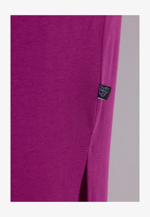 Lähikuva violetista kankaasta, jossa on sivusauma ja pieni musta vaatetunniste lähellä sivuhalkiota.