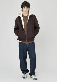 Veste zippée marron avec doublure de capuche crème, associée à une chemise marron et un pantalon en denim bleu. Le look est complété par des baskets blanches.