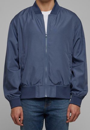 Man wearing a navy blue zip-up bomber jacket with ribbed cuffs and waistband over a white shirt and blue jeans, standing against gray background.