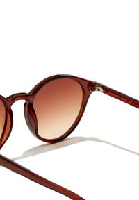 Pyöreät aurinkolasit, joissa on läpinäkyvä burgundin värinen kehys, liukuvärjätyt ruskeat linssit ja hopeiset yksityiskohdat. "Suunniteltu Espanjassa" kaiverrettuna temppeleihin.